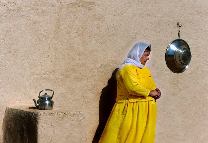 Yezidi woman in yellow dress and white scarf stands against the wall of a building, staring off screen